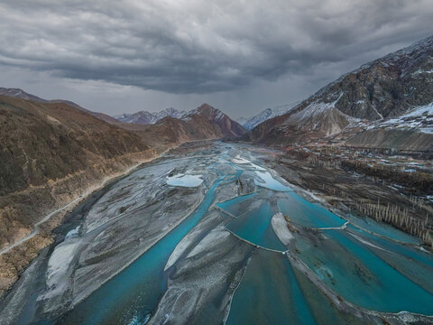 Aerial view of a braided turquoise river flowing through a mountain valley with snow-capped peaks under a cloudy sky in Chitral, Khyber Pakhtunkhwa, Pakistan.