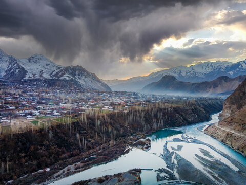 Aerial view of Boni town in Upper Chitral with a turquoise river flowing through a valley surrounded by snow-capped mountains under storm clouds Chitral, Khyber Pakhtunkhwa, Pakistan.