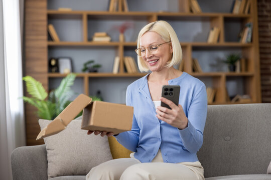 Smiling blonde woman on a couch unboxing a delivered parcel while checking her smartphone, enjoying convenient online shopping, successful delivery and happy customer satisfaction