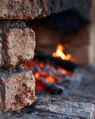 Fototapeta premium Warm fire glows within rustic stone fireplace. Embers flicker softly, casting amber light. Brick edges show weathered, textured surfaces. Cozy, inviting atmosphere radiates from hearth