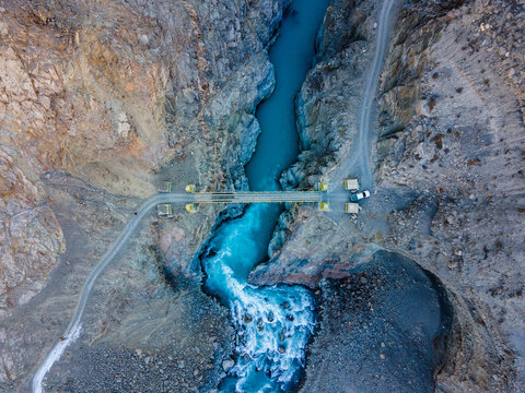 Aerial view of the Boni bridge spanning a turquoise river flowing through a deep rocky canyon with steep cliffs and a winding dirt road in Chitral, Khyber Pakhtunkhwa, Pakistan.