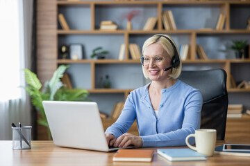 Fototapeta premium Smiling mature woman wearing headphones with microphone actively engaging in remote work, participating in an online meeting, or providing customer support from her home office