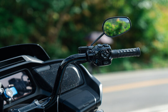 Close up of motorcycle handlebar with rearview mirror, showcasing grip and control buttons against blurred outdoor background