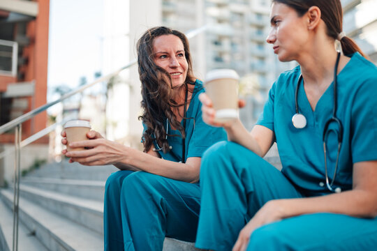 Healthcare workers in teal scrubs sitting on stairs, enjoying coffee during break, discussing work outdoors