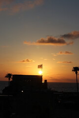 Naklejka premium Caesarea, Israel - JUNE 2, 2026: A flag waves atop a silhouetted building against a vivid orange sunset over the ocean, with palm trees framing the scene.