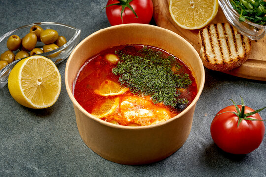 Traditional red hodgepodge solyanka soup with lemon, olives and fresh dill in craft paper bowl, high angle view of hot vegan or vegetarian soup on dark background, eco takeaway food delivery