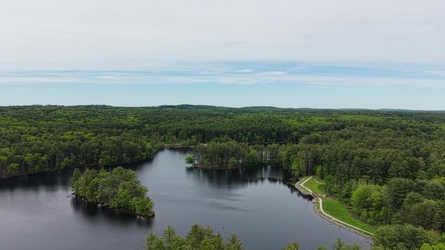 Drone view of dark lake water and small islands surrounded by dense green woodland under cloudy sky in Harold Parker State Forest, Massachusetts, US