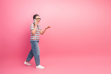 Senior woman talking on smartphone with cheerful expression against pink background, wearing casual...