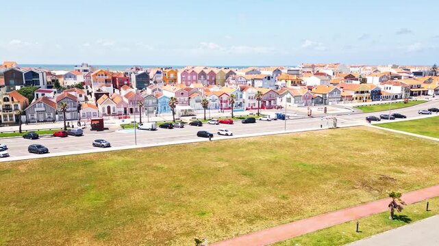 Cars driving past colorful houses beside a grassy open area on a sunny day