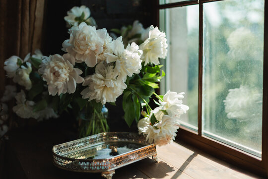 Ornate silver mirror tray with white fluffy feathers on a wooden windowsill beside a bouquet of peonies.