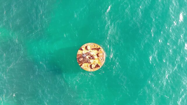 Aerial view of a buoy floating in turquoise sea water, showcasing the buoys circular shape and rusted texture, surrounded by clear ocean waves and reflections of sunlight. Ocean navigation safety