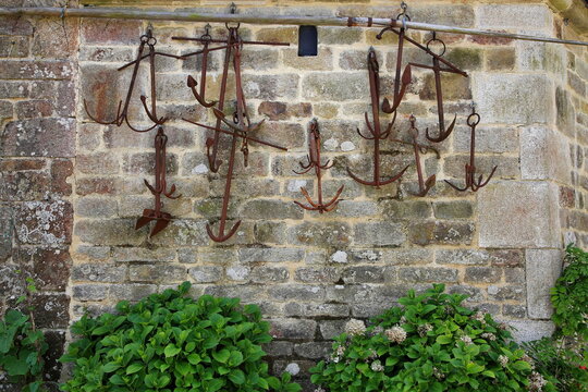 Mehrere rostige, historische Schiffsanker h&auml;ngen als Dekoration an einer alten Natursteinmauer in Locronan. Gr&uuml;ne Hortensienb&uuml;sche s&auml;umen den unteren Bildrand vor dem Mauerwerk.