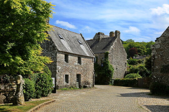 Das Bild zeigt eine charmante, kopfsteingepflasterte Stra&szlig;e in Locronan mit historischen Steinh&auml;usern unter blauem Himmel, umgeben von &uuml;ppigem Gr&uuml;n und einem alten Steinkreuz im Vordergrund.