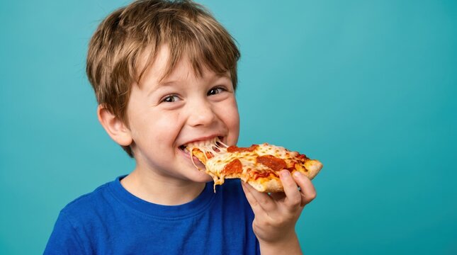 A young boy takes a big bite from a slice of pizza while smiling. He wears a blue shirt. The background is a flat blue color. His expression shows joy and delight in the moment
