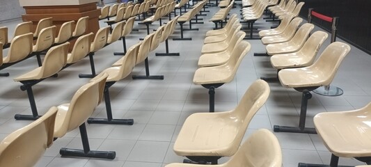 A long row of empty metal and plastic chairs is arranged neatly along the wall of a brightly lit hospital waiting room hallway. © Chawaphan