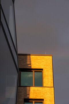 A brick facade holds one window glow at dusk, where shadow falls across an apartment corner and modern sky tightens the quiet edge