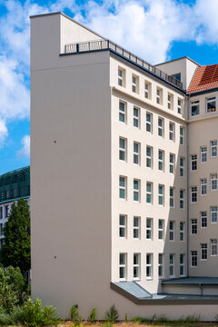 A white building facade rises under sky and clouds, with trees beside residential urban architecture and simple windows across the wall