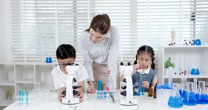 Asian female teacher instructing boy how to use microscope while young girl preparing experiment tools during chemistry class in primary school science room with beakers and test tubes on desk