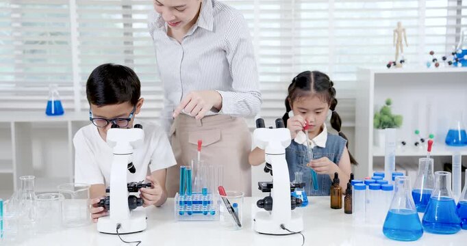 Asian girl transferring blue liquid with pipette during primary school science experiment while boy focusing on microscope and female teacher assisting both students in hands-on chemistry learning