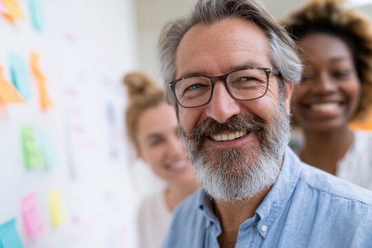Happy man smiles at camera with glasses and beard. Behind him, colleagues smile, adding warmth to scene. Colorful sticky notes dot white wall, signaling teamwork. Ideal for business, collaboration