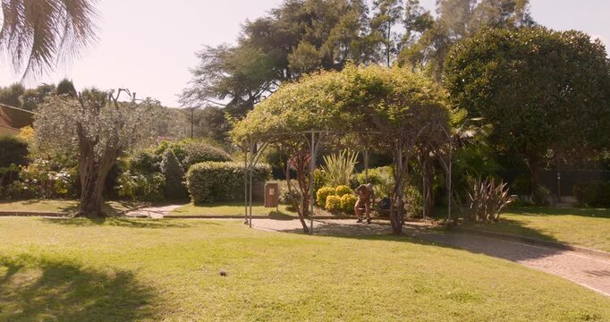 Man Walks And Sits On Bench Under Rustic Gazebo Covered In Climbing Foliage In Saint Jean Cap Ferrat, France. wide shot