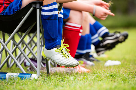Youth soccer players wait their turn on the sidelines of a game in folding chairs, a kid in silver cleats with neon laces sitting beside teammates in blue and red striped team socks