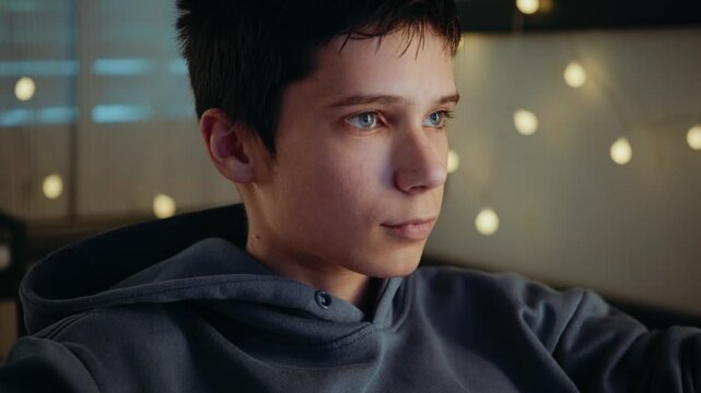 Close-up of a teenage boy's focused face as he plays a video game on his computer at night. His hand is on a gaming mouse.