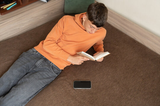 Teenage boy reading book while relaxing on sofa with smartphone lying nearby. Concept of digital balance, healthy screen habits, offline activities, education and mindful leisure time.