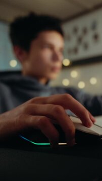 Close-up of a hand on a computer mouse with RGB lights. A teenage boy is playing a video game in his room at night. Vertical video.