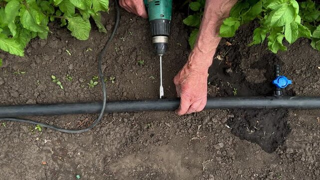 Horizontal video close-up of hands repairing a black plastic garden irrigation pipe with a cordless screwdriver tool.