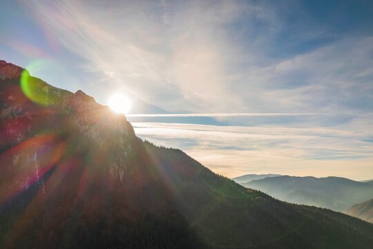 Sunlight gently breaks over the rugged mountain peaks, casting warm light across lush valleys below. The clear blue sky showcases soft clouds as day awakens