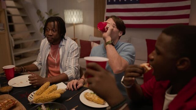 Dolly out of diverse young adults celebrating Independence Day at home, dancing and raising red cups for toast in front of American flag. Footage is ideal for holiday marketing and diversity campaigns