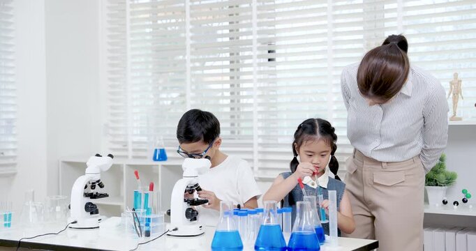 Asian elementary students doing science experiment as girl pours blue liquid into cylinder and teacher observes while boy continues microscope work during practical STEM class in modern classroom