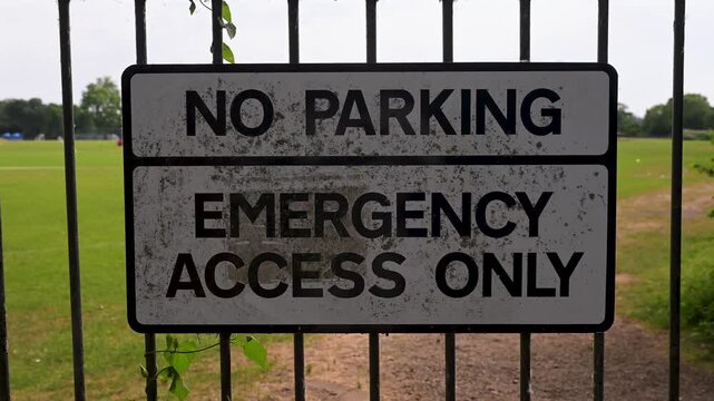 A weathered 'No Parking, Emergency Access Only' sign is mounted on a metal fence, set against a lush green field. Useful for safety and regulation themes.