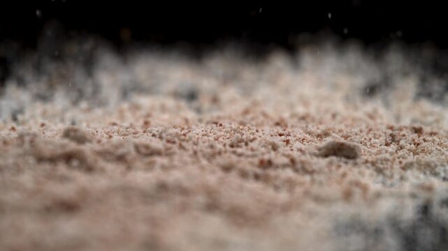 Fine powder flour or makeup particles fall and spread across black shimmering mirror finish background, dramatic macro slow motion