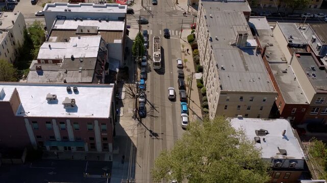 Aerial drone view of a West Philadelphia urban street intersection with flat-roofed commercial buildings, parked cars, and a city bus in traffic below.