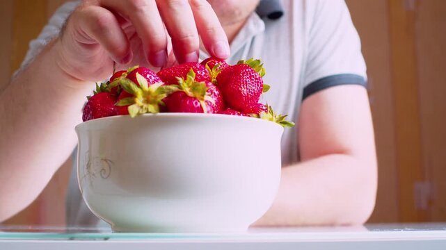 Low angle close-up of man's hands eating ripe strawberries from a bowl, blurred silhouette background. Shallow depth of field, natural window light.Healthy nutrition -organic food. 