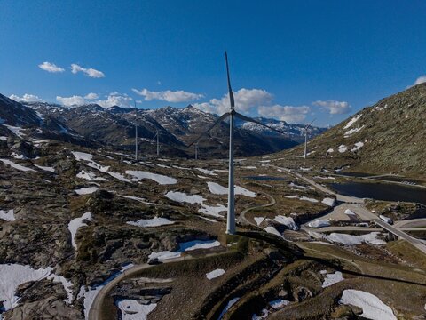 Aerial view of the Gotthard Wind Farm with wind turbines standing on rocky mountain slopes with patches of snow under a clear blue sky in Airolo, Ticino, Switzerland.