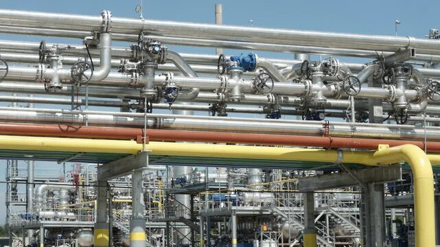 Medium shot of an overhead gas distribution header showing complex pipe networks interconnecting multiple industrial plant units beneath a clear sky. Closeup of labeled pipeline