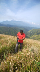 Fototapeta premium Man in a red shirt holding a camera while hiking through a golden meadow, with rolling green hills and a distant mountain peak beneath a clear blue sky.