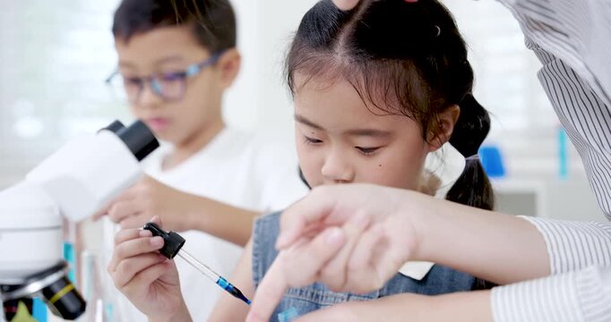 Young Asian girl concentrating while learning to adjust lab tool under teacher's guidance in elementary school science room during chemistry experiment with lab instruments on table around children
