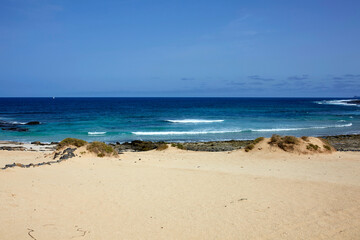 Fototapeta premium Sand dunes with shrubs opening to turquoise ocean with sailboat on horizon, La Graciosa