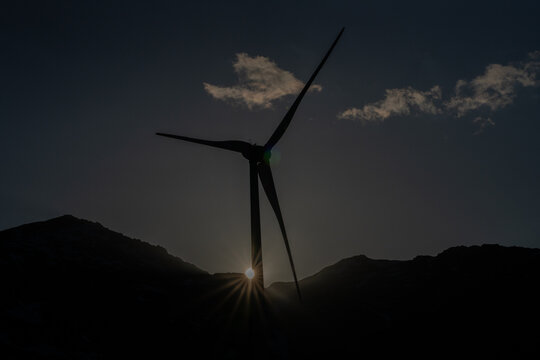 Aerial view of the silhouette of a wind turbine at the Gotthard Wind Farm with a sunstar effect over mountain ridges in Airolo, Ticino, Switzerland.