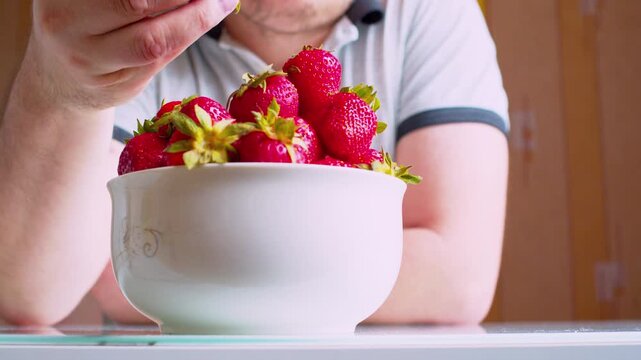 Low angle close-up.Man eating fresh strawberries from a bowl. Blurred man silhouette. Shallow DoF, natural window light. Healthy nutrition, organic food, vitamins and clean lifestyle.
