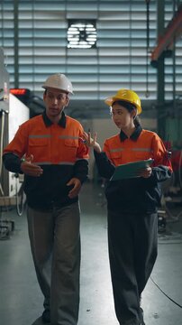 Factory Walkthrough: Two industrial workers in safety vests and hard hats walk through a factory, discussing plans on a clipboard, highlighting collaboration and the manufacturing environment.