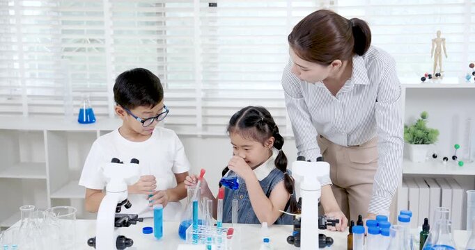 Asian female teacher overseeing young Asian boy and girl performing experiment with beaker and blue liquid while standing beside microscope during creative science class in modern school laboratory