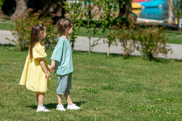 Fototapeta premium Two children, girl in yellow dress and a boy, stand hand in hand on a grassy area in a park, surrounded by trees and colorful water vehicles in the background. Happy childhood concept. Summer vacation