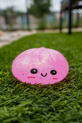 Cute pink glitter squishy toy resembling a smiling bun resting on green grass, playful kawaii character photographed outdoors in vertical format © Juli