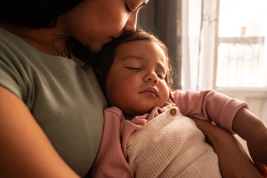 Latin mother cuddling sleeping baby girl by window light
