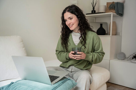 Woman on sofa using laptop with warm drink at home